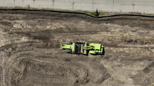 Top-down aerial view of a self-propelled earth scraper cutting and loading soil during site preparation, filling its bowl while grading a construction site.
