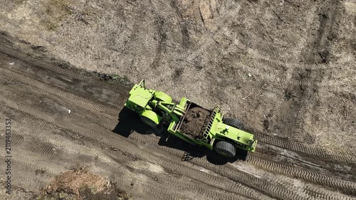 Earth scraper transports a full load of soil along a dirt construction track, with loose dirt spilling from the overloaded bowl, filmed from a rear 45-degree aerial drone perspective.
