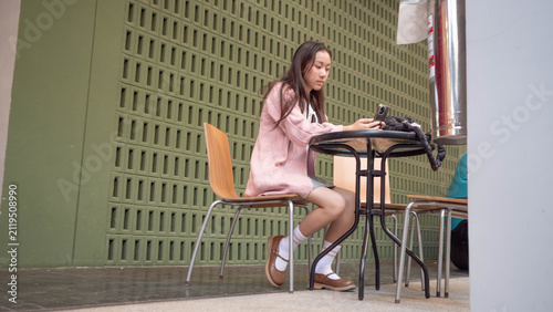 Young Asian woman writing in notebook and using mobile phone at a table outdoors. Remote study, productivity, and lifestyle stock photo.