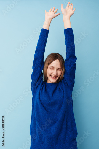 Fotografie Cheerful woman in a blue sweater raises arms in celebration against a bright blue studio backdrop, conveying joy, energy, and positive mood, with sparkling eyes and upbeat vibe