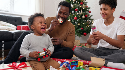 A playful toddler plays with a toy train near father and brother beside a Christmas tree for joyful holiday family promotional imagery