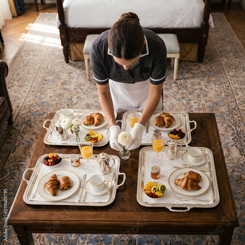 High angle photo of a hotel waitress in a uniform and white gloves arranging flowers and breakfast trays on a wooden table in a luxury hotel room.