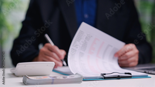 Male accountant reviews financial document, signs clipboard paper, calculates tax budget, manages cash stack, working alone inside office desk, analyzing business investment profit.
