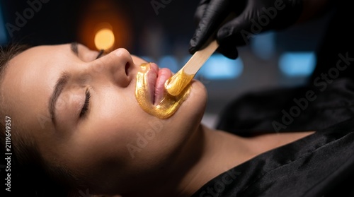 Close up shot of woman receiving facial treatment featuring golden lip mask application, aesthetician wooden spatula and luxury cosmetic care, illustrating skin rejuvenation and spa wellness