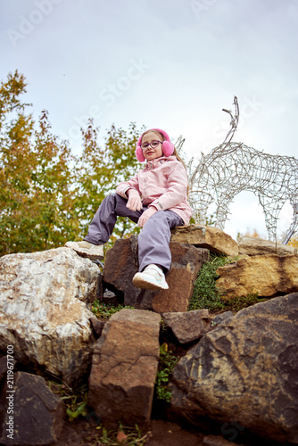 A smiling girl wearing pink earmuffs, glasses, and a light jacket sits on large rocks beside a decorative wire deer sculpture in an autumn park. The low-angle portrait captures an adventurous moment