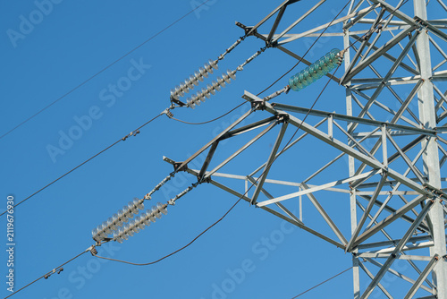 High voltage transmission tower with ceramic insulators, metal lattice structure, and suspended power conductors against clear blue sky in close-up outdoor industrial view
