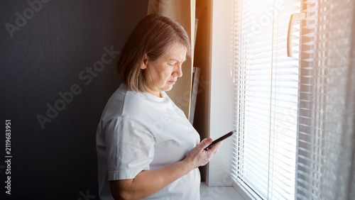 Senior woman standing near a sunlit window with blinds, looking at her smartphone with a serious expression, connecting with technology and the digital world from home