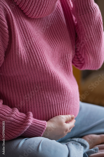 Pregnant woman cradling her belly while seated indoors, wearing a cozy knitted sweater, soft natural light