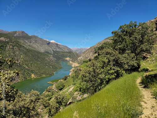 Fotografía Grassy trail traces a sunlit hillside above a winding blue lake, snow-dusted pea