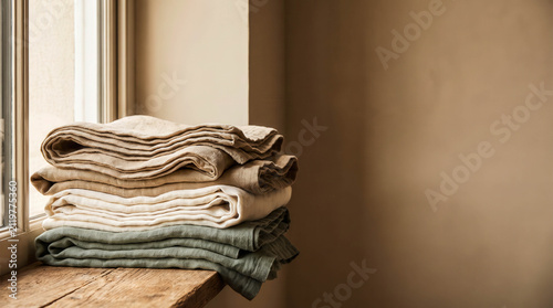 Stack of folded linen textiles in soft neutral colors on a rustic wooden windowsill, illuminated by warm natural light for a cozy home interior.