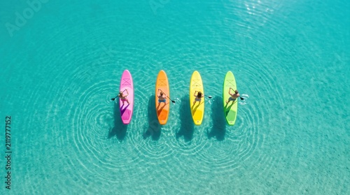 An aerial view of four colorful paddleboards arranged in a row floating on crystal clear turquoise water, with paddlers casting shadows below.