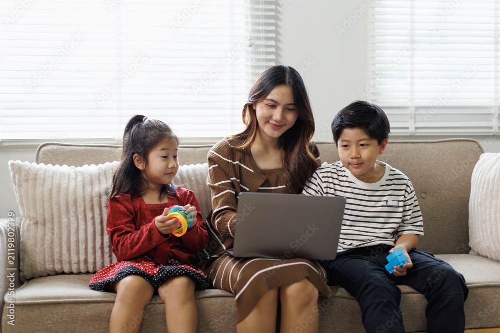 Fototapeta premium Mother and two children sitting on a sofa in a living room, using a laptop together for online learning, enjoying family time, and building strong educational bonds at home