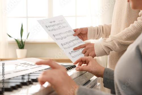Teenage boy with note sheet and music teacher playing synthesizer in classroo...