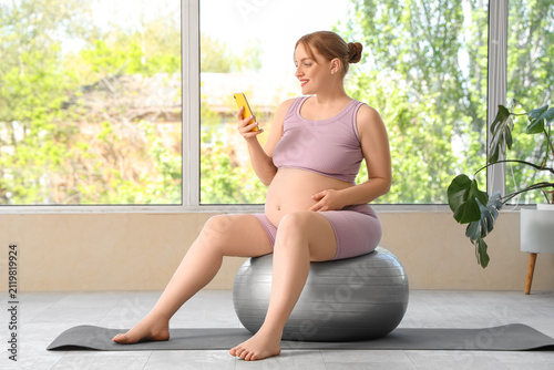 Sporty pregnant woman using mobile phone on fitball near window at gym