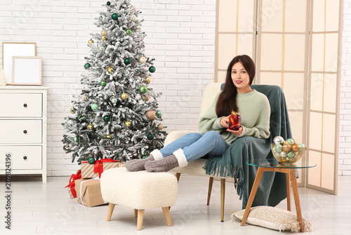 Beautiful young happy Asian woman with cup of hot mulled wine sitting on armc...