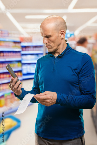Картината върху платно White man comparing receipt and smartphone in supermarket aisle, focused express