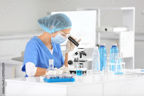 Female young scientist with microscope and professional glassware examining w...