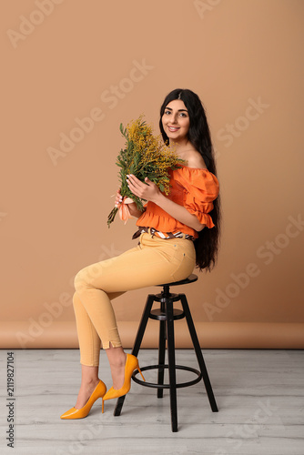 Beautiful young woman with bouquet of mimosa flowers sitting on stool near br...