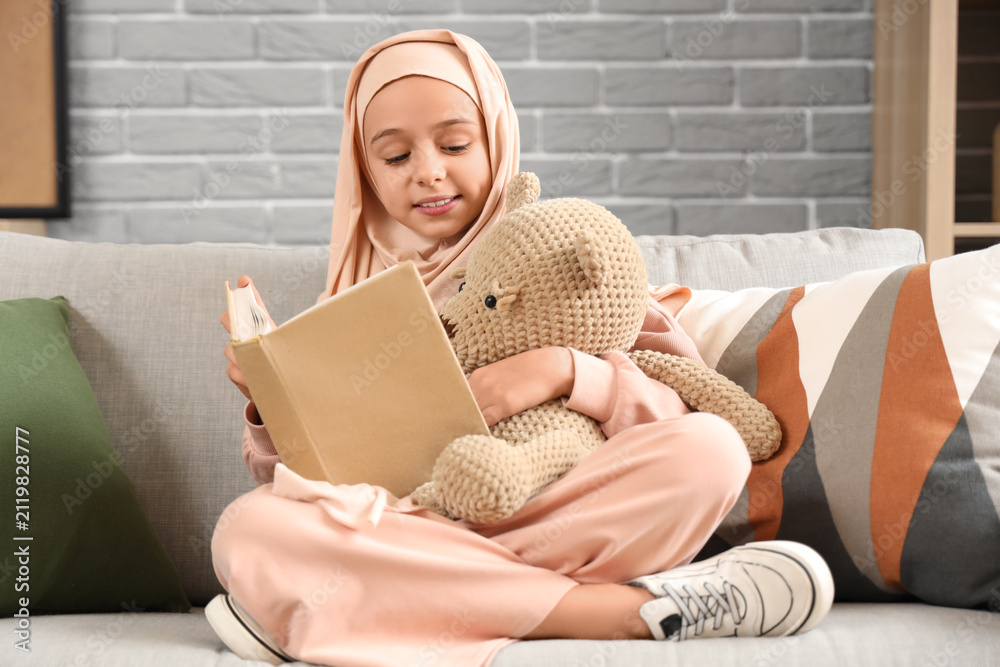 Fototapeta premium Little Muslim girl in hijab with teddy bear reading book at home