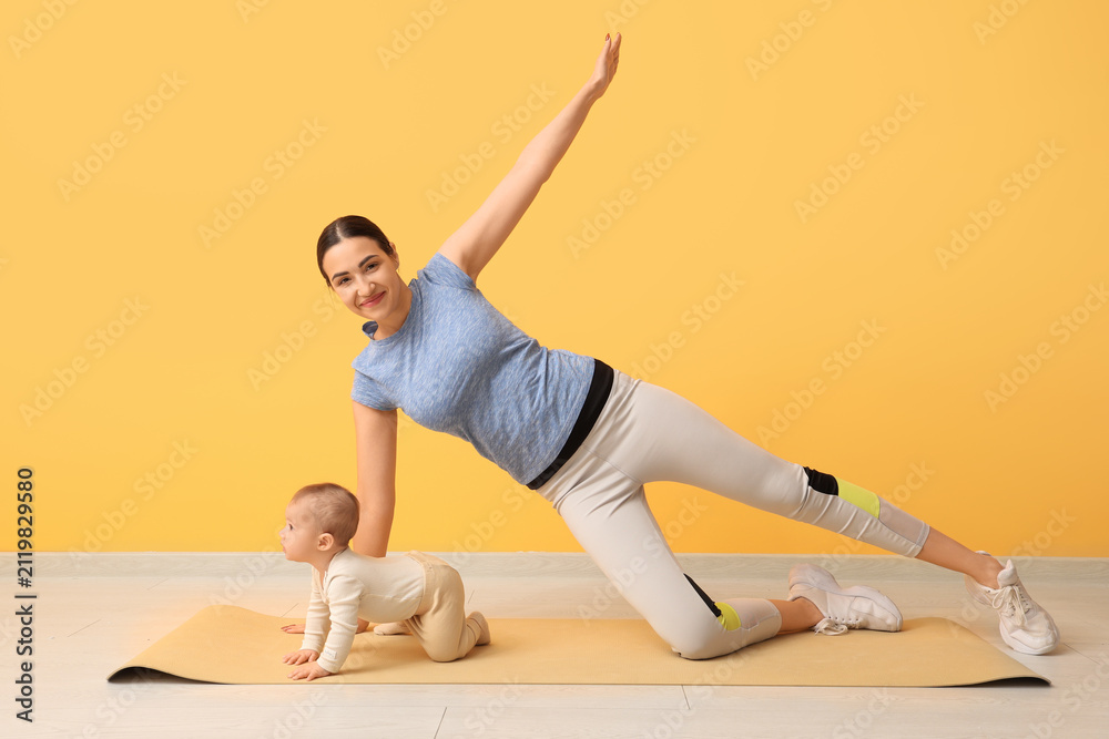 Fototapeta premium Beautiful young sporty woman with her little baby practicing yoga near yellow wall