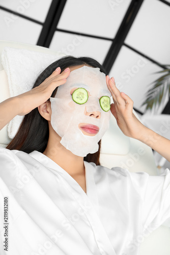 Young Asian woman with cucumber slices and facial sheet mask relaxing in spa ...