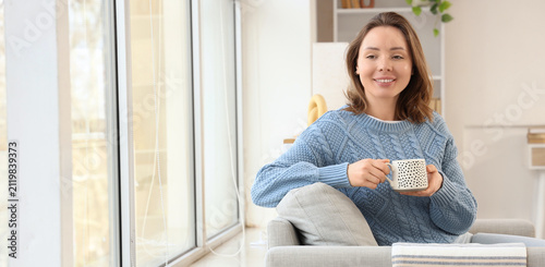 Smiling young woman in a blue knitted sweater relaxing on a sofa with a cup o...