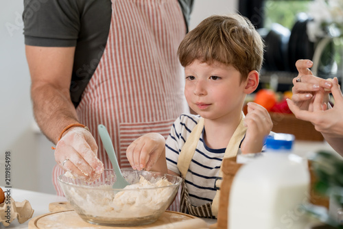 Cuadro en lienzo Caucasian young boy pinches dough inside bowl while father handles floury mixture and mother assists nearby, illustrating sensory learning, family teamwork, parenting, and homemade pastry preparation