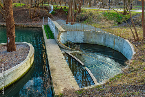 A concrete spillway for a small river into an underground sewer. A spiral hydraulic structure on a city canal in a park. An engineering system for directing water flow among trees