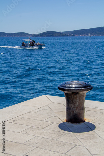 Metal bollard on a pier overlooking blue water of the Mediterranean Sea, with a small motorboat passing in the distance