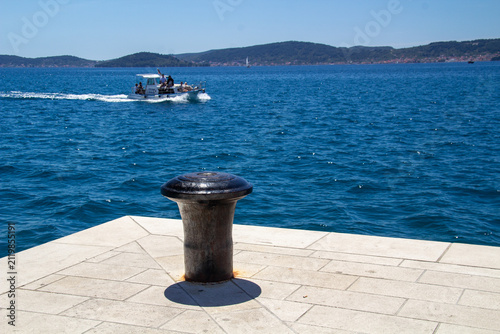 Metal bollard on a pier overlooking blue water of the Mediterranean Sea, with a small motorboat passing in the distance