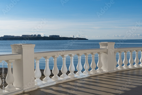 Waterfront Gelendzhik promenade with white balustrade, overlooking calm body of water under clear blue sky, with distant buildings and cranes visible on horizon.