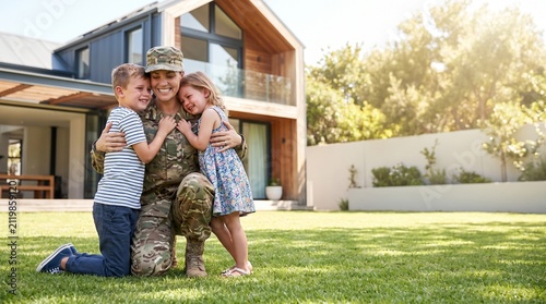 Proud female military soldier kneeling on grass and hugging her small children during a happy homecoming reunion