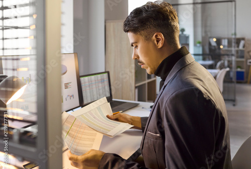 Businessman analyst in office reviews accounting documents. He is a manager auditing charts beside a monitor and laptop, checking audit figures. Clear concept of finance and business analysis.