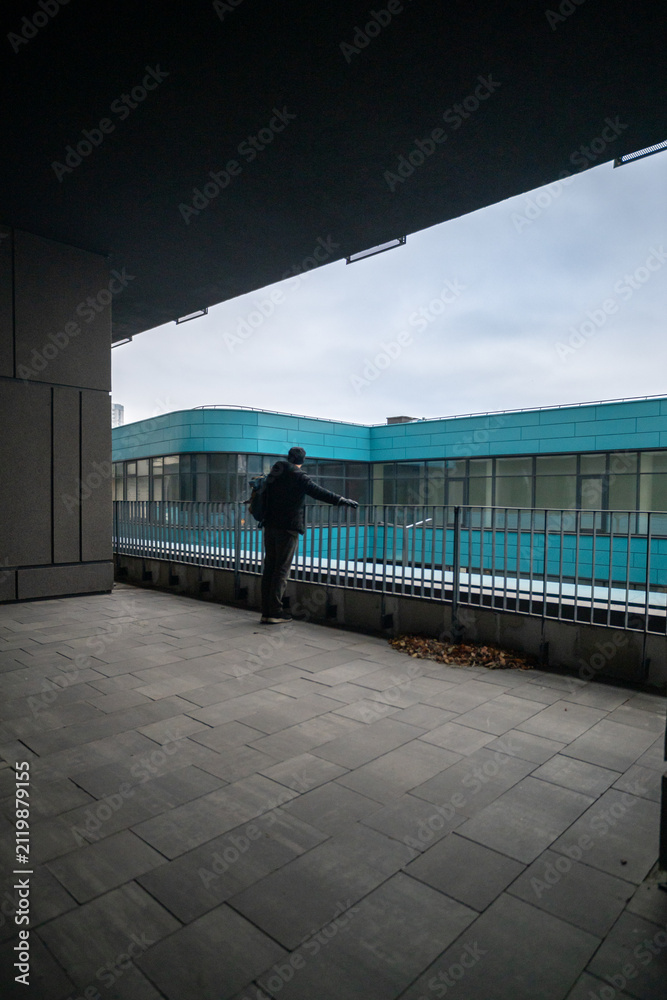 Obraz premium Man Standing on Modern Balcony Looking at Turquoise Office Building Architecture