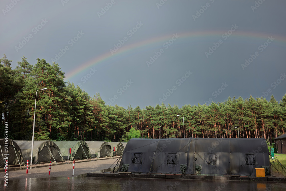 Obraz premium A rainbow arcs over rows of green army tents beside a pine forest after a rain shower at a military camp.