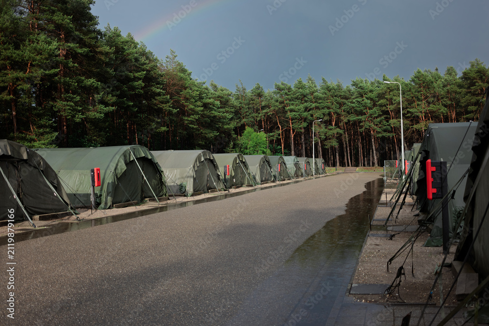 Obraz premium A rainbow rises over rows of green army tents and a pine forest after a summer rain shower at a military camp.