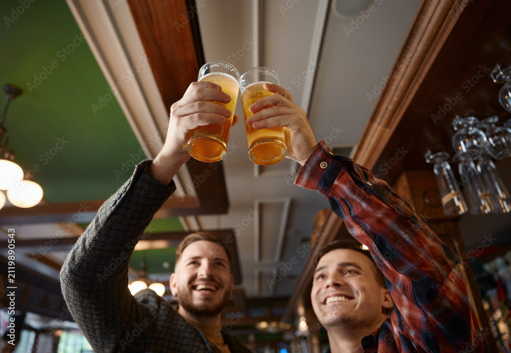 Obraz premium Two cheerful caucasian males celebrate with raised beer glasses and bright pub reflections