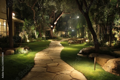 Elegant stone path winds through a lush, illuminated garden at night. Ideal for luxury living, landscape design, or tranquil evening concepts.