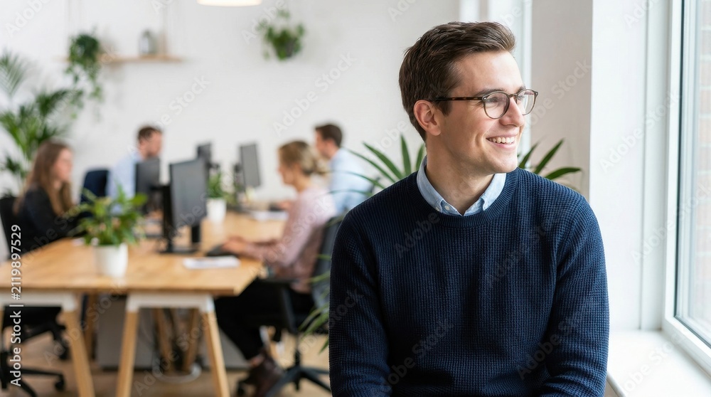 Fototapeta premium Happy Professional Man With Glasses Smiling While Looking Out A Bright Modern Office Window With Diverse Colleagues Working Collaboratively Behind Him