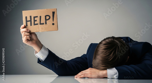 Overworked businessman slumped over desk, holding up a cardboard sign saying HELP! symbolizing stress and burnout in business.