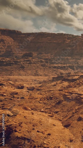 A wide view shows a rocky desert landscape with orange and brown tones. The terrain is uneven with large stones and cliffs. Clouds linger above, casting shadows over the ground.