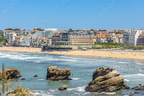 Large rock outcrops rising from the ocean water along the crowded sandy shore of Biarritz on a bright day