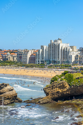 Rock outcrops in the Atlantic Ocean with coastal apartments and the sandy beach visible in the background under a clear sky in Biarritz, France