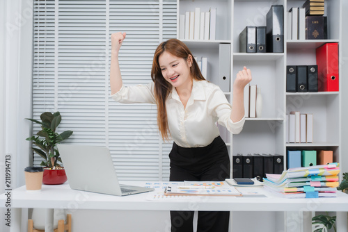 Принти на полотні Celebrating Success at Desk: A young businesswoman cheers with joy and triumph as she achieves a goal at her desk, surrounded by office supplies and a laptop