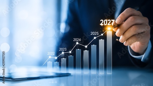 A business person touching a digital bar chart showing financial growth with increasing yearly progress from 2023 to 2027 in a professional office environment with bright blue light effects
