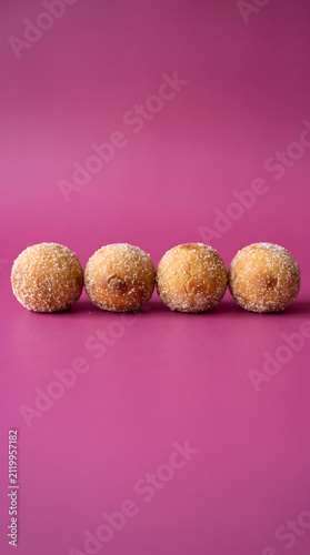 Four freshly made sugarcoated doughnut holes lined up on a vibrant pink surface