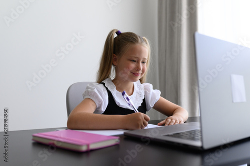 Принти на полотні Diligent girl writing in copybook and smiling at laptop screen during online les