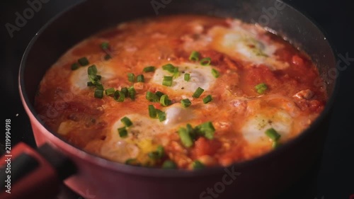A person prepares a dish by cooking eggs shakshuka in a tomato sauce, stirring occasionally. Fresh green herbs are added to enhance the dish's flavor and presentation
