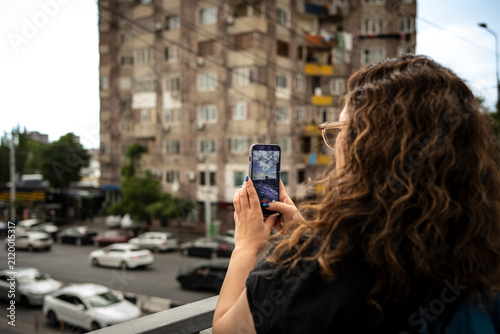 Woman taking smartphone photo of urban city street from balcony, modern travel technology concept