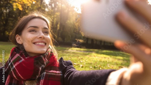 Young woman taking a selfie with a smartphone in a sunlit autumn park, wearing a red plaid scarf and smiling during a beautiful golden hour walk